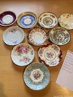 Full view of 11 saucers arranged on a table showing various floral and ornate patterns with different color schemes including blue, maroon, cream, and turquoise backgrounds.