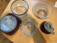 Multiple clear glass bowls with different textures and one square patterned glass bowl arranged on table alongside a stack of small nested amber glass bowls and a stack of amber glass plates.