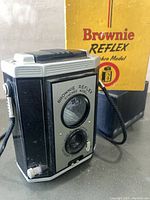 Photo of the Kodak Brownie Reflex Synchro Model camera from an angle highlighting front and side with black strap attached. Original yellow box visible in background.