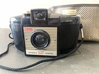 Front view of Kodak Brownie 127 vintage camera with visible lens, shutter button, and wrist strap, placed on surface with carry case behind