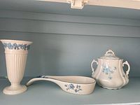 Three blue and white china pieces on shelf: Wedgwood vase, spoon holder, and double-handled jar with lid.