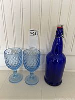 Two blue glass goblets with diamond pattern and matching stems and bases, next to a tall blue glass bottle with swing top closure.
