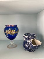 Full view of blue glass stemmed vase with hand painted floral decoration and blue and white shaving mug on a shelf.