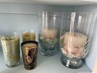Photo of the two large glass vases with coral etching, each containing a white pillar candle with seashells and glass beads, alongside four decorative candles including two green with beads, one black and gold, and one cream candle