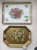 Photo showing both metal trays: a rectangular white tray with colorful floral center and border detailing, and a gold octagonal tray with white flowers and lattice cut-outs.