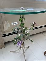 Side angle showing metal pedestal base with painted floral and vine decorations in multiple colors and round glass top with crack.