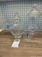 Four clear glass vessels displayed on a counter with tile background: large apothecary jar, medium floral etched jar, round cornflower vase, and small cylindrical vase.