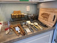 Photo shows the wood bamboo cutting board with pig design, metal utensil rack, spice rack with labeled boxes, glass casserole dishes, and various cooking utensils including wooden spoons, ladle, spatula, tongs, mesh strainers, and scoops.
