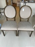 Pair of dining chairs with oval cushioned backs and upholstered seats shown from front, placed on light-colored floor.