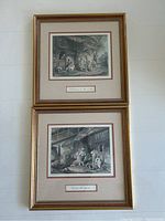 Pair of framed French prints stacked vertically against a white wall, each with gold-toned frame and beige/red mats, featuring outdoor village scenes.