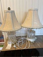 Pair of glass base table lamps with beige fabric shades on wooden surface, one lamp missing finial.