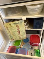 Plastic containers and kitchen items organized inside a drawer with a metal wall decoration leaning against the inside side of the drawer.