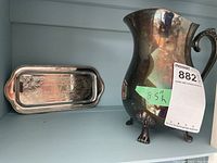 Silver plated claw foot pitcher alongside small silver plated tray on display shelf.