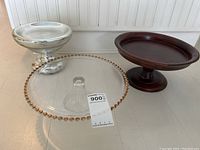 Photo of three platters: clear glass cake plate with gold hobnail edge, dark wooden pedestal plate, silver-colored metal pedestal candy dish.