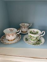 Three Royal Albert teacups and saucers arranged on a shelf: a pair of 50th Anniversary cups with gold floral motif, and one Lily of the Valley cup with 'May' inscription.