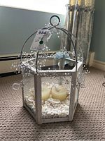 Front angled view of whitewashed hexagonal candle lantern showing glass panels, candles and seashells inside, with decorative bead garland on top.