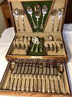 Complete flatware set arranged in open wooden box with upper section holding serving pieces and lower section holding knives with cream handles.