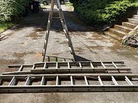 Three ladders displayed on a driveway: wooden step ladder standing upright, two aluminum extension ladders lying flat.
