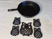 Cast iron skillet and five owl-shaped cast iron trivets arranged flat showing detail and shape