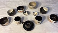 Top view of 11 ceramic dishes including small teacups, small bowls and a saucer arranged on white cloth.