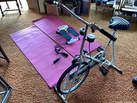 White stationary exercise bike with black pedal and seat placed over carpet next to pink yoga mat and stepper.