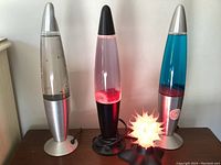 Front view of three lava lamps with a small red decorative lamp. Blue lava lamp shows red wax and blue liquid; clear lamp has some floating particles; pink wax lava lamp illuminated.