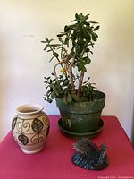 Jade plant in green plastic pot, ceramic vase with leaf pattern, and plastic crashing wave figurine displayed on a pink cloth against a plain wall