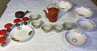 Full view of Japanese dishes including five small tea cups with red lids, a white plate, ceramic bowls, and two teapots on a white cloth.