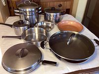 Overview of pots and pans on table, including stainless steel pots, copper frying pan, black wok with glass lid