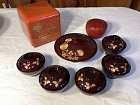 Full set showing six dark brown lacquered resin bowls with lids and matching leaf pattern, circular lacquer plate, red lacquered container, and stacked red layered box