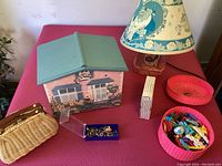 Photo showing house-shaped jewelry box, Beauty and the Beast lamp and book, small wicker handbag, blue container with rings, and pink bowls with hair accessories on a red surface.