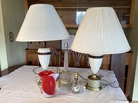 Four vintage lamps and a red-white glass pitcher displayed on a table with lamp shades on the ceramic lamps.