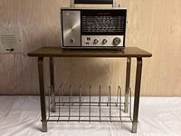 Front view of vintage record table with laminate top and metal legs, featuring metal rack for holding vinyl records. On top is a vintage black and silver Sylvania radio with dials and tuning display.