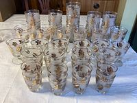 Front view of a large assortment of vintage Libby glassware with gold leaf and striped gold rim decorations arranged on a table.