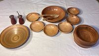 Set of wooden bowls arranged on a white surface, showing two larger bowls, six smaller bowls, wooden serving utensils, and salt and pepper shakers.