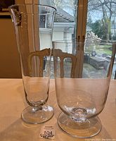 Two large clear glass decorative pieces, one tall flared vase and one shorter wide-mouthed vase displayed on a table with window background.