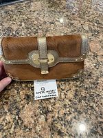 Front view of brown calf hair clutch with gold metal trim, clasp, and grommet detail placed on granite countertop with sale auction tag.