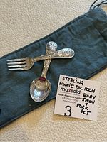 Two child-sized sterling silver utensils, a spoon and fork, displayed crossed on a dark fabric pouch. Spoon has an embossed Winnie the Pooh figure on the handle, while fork has Tigger.