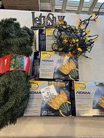 Photo showing the 9-foot green artificial garland wrapped with a label, four unopened packages of NOMA LED gold string lights (70 lights each), and a 'let it snow' metal decorative sign. Lights are yellow and shaped like tulip flowers.