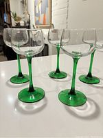 Front angle view of five vintage wine glasses with clear bowls and green stems and bases grouped on a white surface.