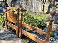 Photo showing the headboard, footboard, and one side rail of a solid wood single bed frame outdoors on stone paving, with garden background