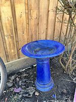 Blue concrete bird bath standing on soil against a wooden fence, showing top view and blue glazed surface with chips and damage.
