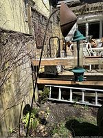 Image of a green hanging bird feeder suspended from a black curved metal stand in an outdoor garden setting near a wooden deck and house wall.