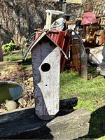 Photo of a rustic wooden birdhouse outdoors, showing front view with circular entrance hole, peaked roof, and hanging wire loop on top.