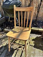 Pine wooden chair with a contoured seat and spindled backrest, shown outdoors on a patio with sunlight casting shadows.