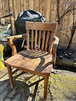 Photo of solid wood armchair showing slatted vertical backrest, curved armrests, contoured seat, and straight legs outdoors on a paved surface with a wooden fence background.