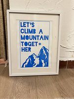 Full view of framed art print leaning against wall on floor. Text 'LET'S CLIMB A MOUNTAIN TOGET+ HER' in blue uppercase with mountain graphic below.