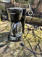 Outdoor photo of a black Black & Decker drip coffee maker with glass carafe and digital display, power cord visible.