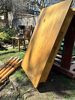 Photo of the natural wood grain tabletop leaning against a structure. Several disassembled round pine legs are visible resting on the ground.