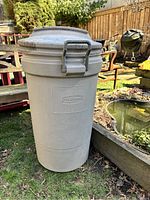 Front view of a tall gray Rubbermaid garbage bin with a lid showing the latch handle and outdoor setting.